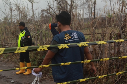 Two individuals stand in an outdoor area with dried vegetation. One person is wearing a uniform with a reflective vest and yellow boots, suggesting an official capacity. The other person, facing away, wears a t-shirt with a logo and web address. Yellow caution tape runs across the scene, indicating a restricted or hazardous area.