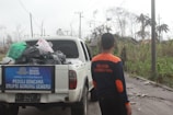 A person wearing a jacket with text on the back stands next to a white pickup truck loaded with bags and supplies. The truck has a banner mentioning disaster relief for the eruption of Mount Semeru. The background shows an outdoor setting with power lines, palm trees, and vegetation affected by the eruption.