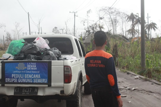 A person wearing a jacket with text on the back stands next to a white pickup truck loaded with bags and supplies. The truck has a banner mentioning disaster relief for the eruption of Mount Semeru. The background shows an outdoor setting with power lines, palm trees, and vegetation affected by the eruption.