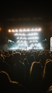 A large crowd is gathered in front of an outdoor concert stage, illuminated by numerous bright lights that cast a glow over the audience. The atmosphere seems vibrant yet intimate, with one performer visible on the stage amidst the haze.