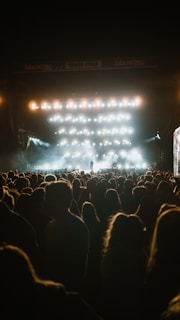 A large crowd is gathered in front of an outdoor concert stage, illuminated by numerous bright lights that cast a glow over the audience. The atmosphere seems vibrant yet intimate, with one performer visible on the stage amidst the haze.