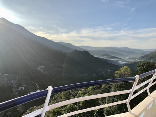 A scenic view from a balcony overlooking lush, green mountains under a clear blue sky. Sunlight streams over the peaks, creating a tranquil atmosphere with mist lingering in the valleys.