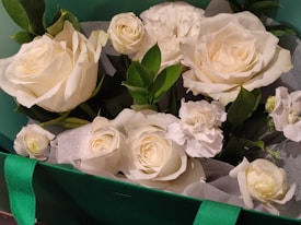 A bouquet of elegant white roses and carnations surrounded by lush green leaves and wrapped in delicate white paper. The flowers are arranged in a decorative manner, emphasizing their freshness and purity.