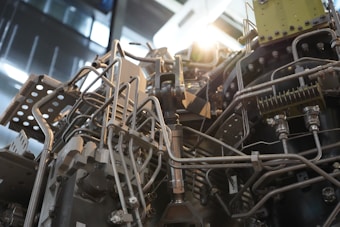A complex assembly of metallic pipes, tubes, and components intertwined, typical of an industrial machine or engine. The structure features various bolts, joints, and fittings, suggesting a mechanized function. The lighting casts a glinting effect on the metal surfaces, highlighting the intricate details of the construction.
