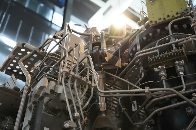 A complex assembly of metallic pipes, tubes, and components intertwined, typical of an industrial machine or engine. The structure features various bolts, joints, and fittings, suggesting a mechanized function. The lighting casts a glinting effect on the metal surfaces, highlighting the intricate details of the construction.