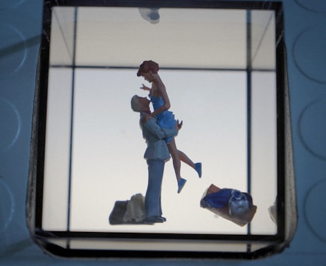 A romantic scene featuring a small figurine of a couple, where one person is lifting the other, creating a sense of affection and joy. The background is minimalistic with soft lighting, and other objects like a bag or clothing piece are visible in the foreground.