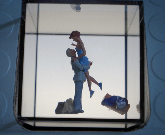 A romantic scene featuring a small figurine of a couple, where one person is lifting the other, creating a sense of affection and joy. The background is minimalistic with soft lighting, and other objects like a bag or clothing piece are visible in the foreground.