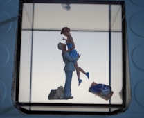 A romantic scene featuring a small figurine of a couple, where one person is lifting the other, creating a sense of affection and joy. The background is minimalistic with soft lighting, and other objects like a bag or clothing piece are visible in the foreground.