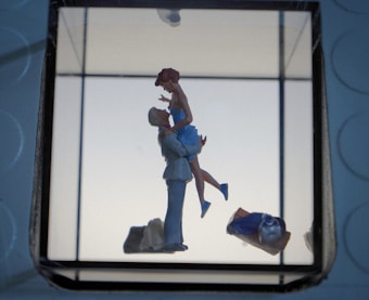 A romantic scene featuring a small figurine of a couple, where one person is lifting the other, creating a sense of affection and joy. The background is minimalistic with soft lighting, and other objects like a bag or clothing piece are visible in the foreground.