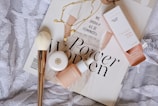 A flat lay composition featuring a magazine with bold 'Power Women' text, a white shirt with feminist text partially visible, a white bristle makeup brush with a gold handle, a necklace with round pendants, and cosmetic products including a small container and a box labeled 'Face Hero'. The items are arranged on a textured, patterned fabric background.