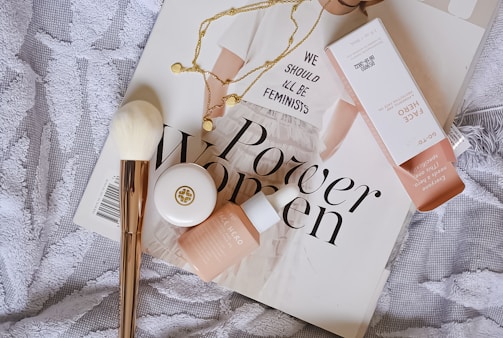 A flat lay composition featuring a magazine with bold 'Power Women' text, a white shirt with feminist text partially visible, a white bristle makeup brush with a gold handle, a necklace with round pendants, and cosmetic products including a small container and a box labeled 'Face Hero'. The items are arranged on a textured, patterned fabric background.