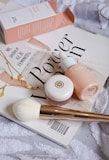 A collection of beauty products arranged artfully on a magazine cover. The setup includes a pink box labeled 'Face Hero', a dropper bottle with a matching color scheme, a small round pot with a golden logo, a white fluffy brush with a golden handle, and a delicate gold necklace with circular charms. The magazine beneath these items displays bold text in a stylish font. The overall composition conveys a sense of elegance and beauty.