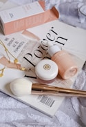 A collection of beauty products arranged artfully on a magazine cover. The setup includes a pink box labeled 'Face Hero', a dropper bottle with a matching color scheme, a small round pot with a golden logo, a white fluffy brush with a golden handle, and a delicate gold necklace with circular charms. The magazine beneath these items displays bold text in a stylish font. The overall composition conveys a sense of elegance and beauty.