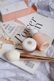 A collection of beauty products arranged artfully on a magazine cover. The setup includes a pink box labeled 'Face Hero', a dropper bottle with a matching color scheme, a small round pot with a golden logo, a white fluffy brush with a golden handle, and a delicate gold necklace with circular charms. The magazine beneath these items displays bold text in a stylish font. The overall composition conveys a sense of elegance and beauty.