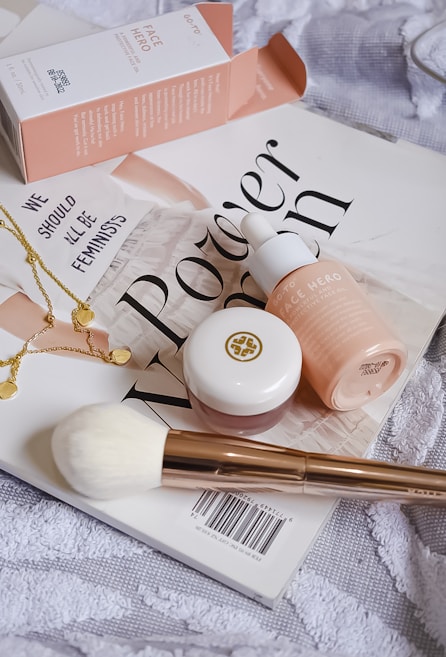 A collection of beauty products arranged artfully on a magazine cover. The setup includes a pink box labeled 'Face Hero', a dropper bottle with a matching color scheme, a small round pot with a golden logo, a white fluffy brush with a golden handle, and a delicate gold necklace with circular charms. The magazine beneath these items displays bold text in a stylish font. The overall composition conveys a sense of elegance and beauty.