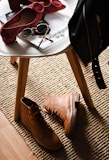 A stylish composition featuring fashion accessories on and around a white round table. There are chic red high-heeled shoes placed on the table next to sunglasses and magazines. On the floor lies a pair of rugged tan boots, and a black leather jacket is draped over the chair. This setup is placed on a textured beige rug, adding warmth to the scene.