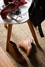 A stylish composition featuring fashion accessories on and around a white round table. There are chic red high-heeled shoes placed on the table next to sunglasses and magazines. On the floor lies a pair of rugged tan boots, and a black leather jacket is draped over the chair. This setup is placed on a textured beige rug, adding warmth to the scene.