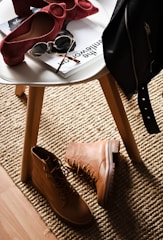 A stylish composition featuring fashion accessories on and around a white round table. There are chic red high-heeled shoes placed on the table next to sunglasses and magazines. On the floor lies a pair of rugged tan boots, and a black leather jacket is draped over the chair. This setup is placed on a textured beige rug, adding warmth to the scene.