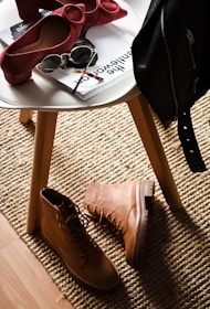 A stylish composition featuring fashion accessories on and around a white round table. There are chic red high-heeled shoes placed on the table next to sunglasses and magazines. On the floor lies a pair of rugged tan boots, and a black leather jacket is draped over the chair. This setup is placed on a textured beige rug, adding warmth to the scene.