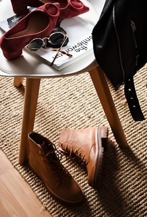 A stylish composition featuring fashion accessories on and around a white round table. There are chic red high-heeled shoes placed on the table next to sunglasses and magazines. On the floor lies a pair of rugged tan boots, and a black leather jacket is draped over the chair. This setup is placed on a textured beige rug, adding warmth to the scene.