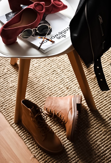 A stylish composition featuring fashion accessories on and around a white round table. There are chic red high-heeled shoes placed on the table next to sunglasses and magazines. On the floor lies a pair of rugged tan boots, and a black leather jacket is draped over the chair. This setup is placed on a textured beige rug, adding warmth to the scene.