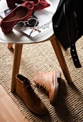 A stylish composition featuring fashion accessories on and around a white round table. There are chic red high-heeled shoes placed on the table next to sunglasses and magazines. On the floor lies a pair of rugged tan boots, and a black leather jacket is draped over the chair. This setup is placed on a textured beige rug, adding warmth to the scene.