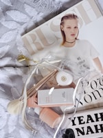 A flat lay featuring a fashion magazine with a model on the cover, a clear toiletry bag containing skincare products and accessories, and a textured white fabric background.
