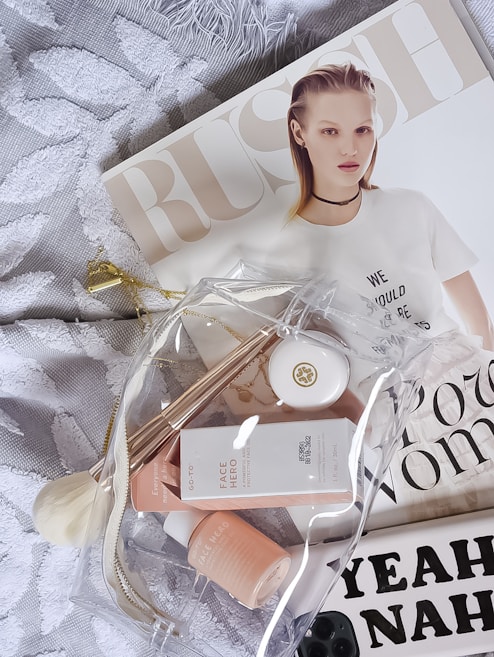 A flat lay featuring a fashion magazine with a model on the cover, a clear toiletry bag containing skincare products and accessories, and a textured white fabric background.