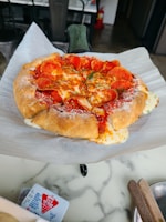A freshly baked pizza with a thick, crispy crust sits on a sheet of parchment paper. The pizza is topped with melted cheese, slices of pepperoni, a sprinkle of herbs, and a dusting of grated cheese. It's placed on a marble countertop, with a fire extinguisher visible in the background. Nearby, there is a red and white container and wooden utensils in the foreground.