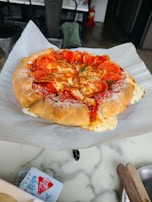 A freshly baked pizza with a thick, crispy crust sits on a sheet of parchment paper. The pizza is topped with melted cheese, slices of pepperoni, a sprinkle of herbs, and a dusting of grated cheese. It's placed on a marble countertop, with a fire extinguisher visible in the background. Nearby, there is a red and white container and wooden utensils in the foreground.