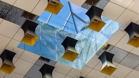 A modern ceiling design with a central skylight composed of glass panes, surrounded by square acoustic panels with a textured surface. Several geometric, black and yellow pendant lights hang at varying angles from the ceiling.