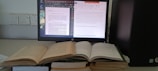 A workspace featuring a computer monitor displaying two digital documents. Next to it, several open books are stacked, and the computer tower is positioned nearby. The monitor shows one document in a PDF reader and another in a word processor.