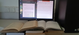 A workspace featuring a computer monitor displaying two digital documents. Next to it, several open books are stacked, and the computer tower is positioned nearby. The monitor shows one document in a PDF reader and another in a word processor.