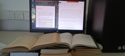 A workspace featuring a computer monitor displaying two digital documents. Next to it, several open books are stacked, and the computer tower is positioned nearby. The monitor shows one document in a PDF reader and another in a word processor.