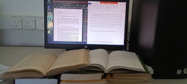 A workspace featuring a computer monitor displaying two digital documents. Next to it, several open books are stacked, and the computer tower is positioned nearby. The monitor shows one document in a PDF reader and another in a word processor.