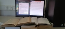 A workspace featuring a computer monitor displaying two digital documents. Next to it, several open books are stacked, and the computer tower is positioned nearby. The monitor shows one document in a PDF reader and another in a word processor.