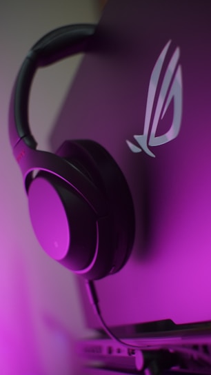 A pair of black headphones is resting on the edge of a laptop, with a logo visible on the laptop lid. The scene is illuminated with a purple hue, creating a modern and stylish ambiance.