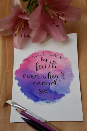 A handwritten quote on a piece of paper with a watercolor background in shades of pink and blue, surrounded by pink lilies and art brushes. The quote reads, 'I will walk by faith even when I cannot see.'