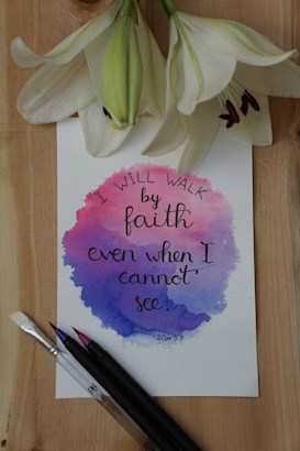 A piece of paper with watercolor design features an inspirational quote in cursive handwriting, surrounded by three white lilies. Paintbrushes with colored tips are placed next to the paper on a wooden surface.