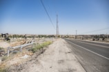 A wide, open road stretches into the distance, bordered by a dry, dusty landscape. On the left side, multiple oil pumps are visible, scattered across the barren terrain. Power lines run along the roadside, while a chain-link fence surrounds the oil field area. The sky is clear and blue.