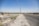 A wide, open road stretches into the distance, bordered by a dry, dusty landscape. On the left side, multiple oil pumps are visible, scattered across the barren terrain. Power lines run along the roadside, while a chain-link fence surrounds the oil field area. The sky is clear and blue.