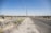 A wide, open road stretches into the distance, bordered by a dry, dusty landscape. On the left side, multiple oil pumps are visible, scattered across the barren terrain. Power lines run along the roadside, while a chain-link fence surrounds the oil field area. The sky is clear and blue.