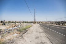 A wide, open road stretches into the distance, bordered by a dry, dusty landscape. On the left side, multiple oil pumps are visible, scattered across the barren terrain. Power lines run along the roadside, while a chain-link fence surrounds the oil field area. The sky is clear and blue.