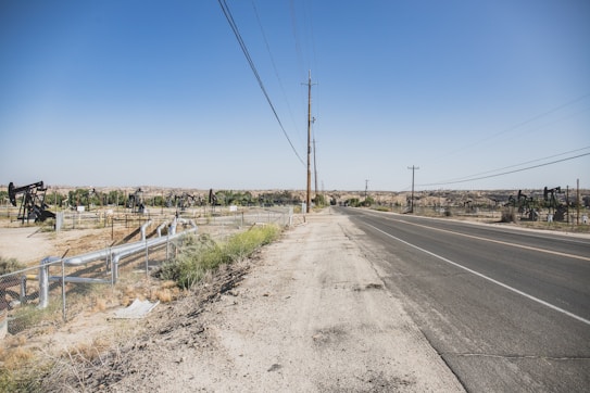A wide, open road stretches into the distance, bordered by a dry, dusty landscape. On the left side, multiple oil pumps are visible, scattered across the barren terrain. Power lines run along the roadside, while a chain-link fence surrounds the oil field area. The sky is clear and blue.