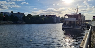 A calm river bordered by buildings and lush green trees on one side and a boat with a sign reading 'Hostel & Lounge' moored to the riverbank on the other. The sky is partly cloudy, and sunlight reflects off the water's surface.