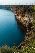 A stunning coastline with steep, rugged cliffs descending into a vast body of deep blue water. At the top of the cliffs, numerous wind turbines stretch across the horizon, harnessing wind energy. Vegetation and foliage are visible in the foreground, adding a touch of greenery.