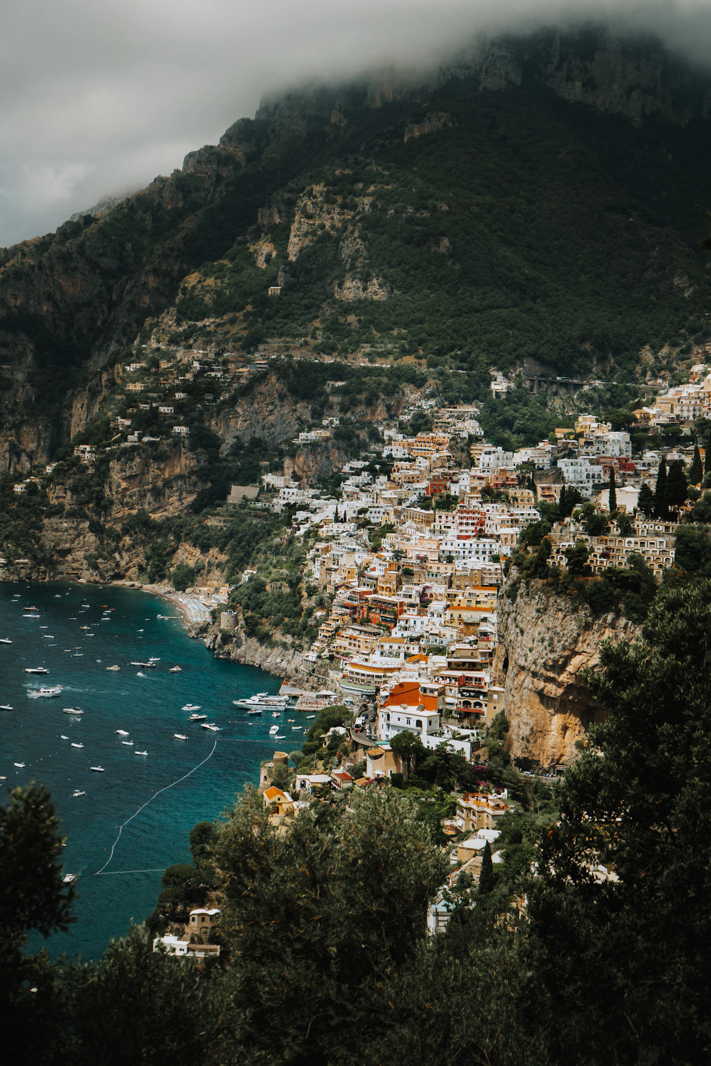 A pastel cliffside town stacked above a turquoise bay, boats moored below and cloud coming over the ridge.