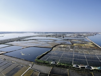Aerial view of salt evaporation ponds located near a coastal area. The ponds form a grid-like pattern with various sections of different colors, ranging from dark blues to light browns and yellows. The horizon is clear and sky is blue with a few distant structures visible along the shoreline.