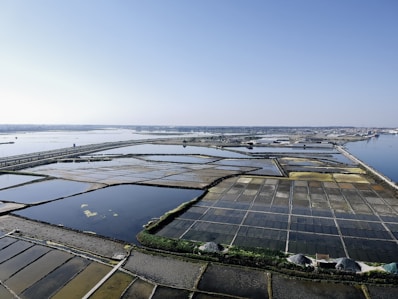 Aerial view of salt evaporation ponds located near a coastal area. The ponds form a grid-like pattern with various sections of different colors, ranging from dark blues to light browns and yellows. The horizon is clear and sky is blue with a few distant structures visible along the shoreline.