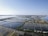 Aerial view of salt evaporation ponds located near a coastal area. The ponds form a grid-like pattern with various sections of different colors, ranging from dark blues to light browns and yellows. The horizon is clear and sky is blue with a few distant structures visible along the shoreline.